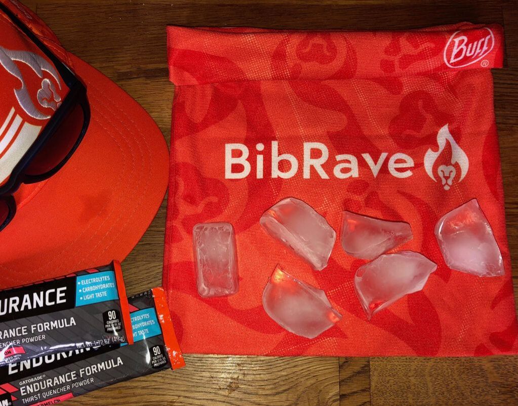 Image of a BibRave classic Buff headwear with 6 ice cubes laying on it. There's a hat and sunglasses on the left side of the photo.